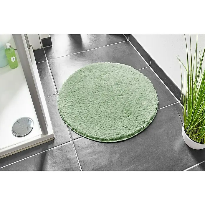 Badteppich Florida - 60 x 60 cm, Salbei, 100% Polyester - Runder, mintgrüner Badteppich im Badezimmer auf dunklen Fliesen, neben einer Dusche und einer Zimmerpflanze.