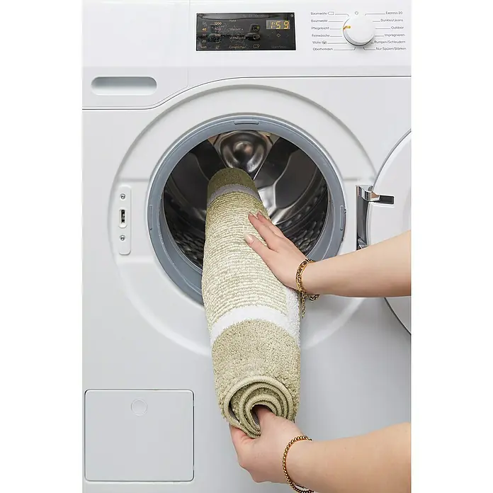 Badteppich Maui - 50 x 80 cm, Beige, 100% Polyester - Eine Person legt eine zusammengerollte, hellgrün-weiß gestreifte Badematte in eine weiße Miele-Waschmaschine.
