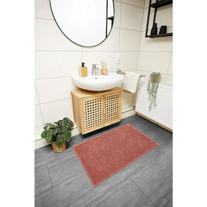 Badteppich Malibu - 60 x 100 cm, Rostrot, 100% Polyester - Badezimmer-Szene mit einer rechteckigen, korallenroten Badematte, die vor einem Waschtisch mit Unterschrank auf dunklen Fliesen liegt.