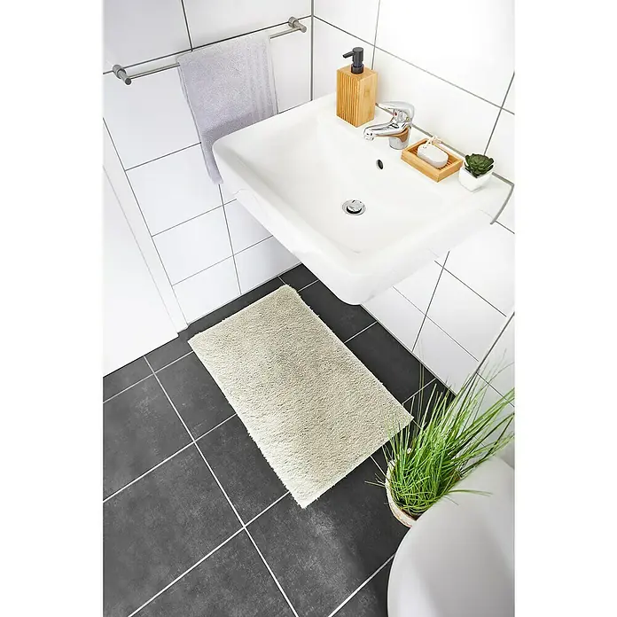 Badteppich Florida - 50 x 80 cm, Beige, 100% Polyester - Badezimmer-Interieur mit einem weißen Waschbecken, einer hellen Badematte auf dunklen Fliesen und einem Handtuchhalter mit Handtuch.