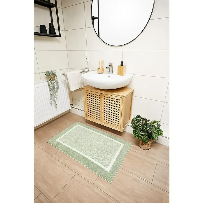 Badteppich Maui - 60 x 100 cm, Salbei, 100% Polyester - Badezimmer-Szene mit einem hellgrünen Badteppich mit weißem Rand auf einem Holzboden, der vor einem Waschtisch mit Rattantüren liegt.