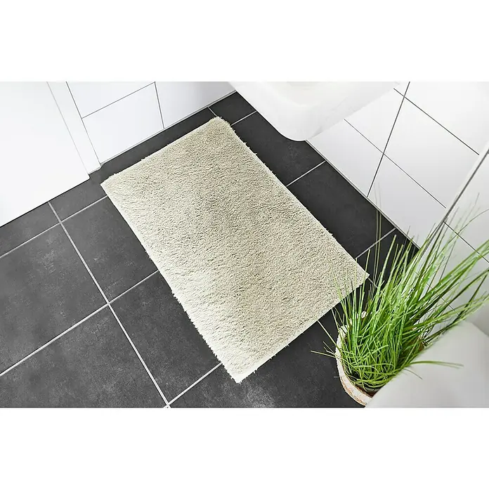 Badteppich Florida - 50 x 80 cm, Beige, 100% Polyester - Ein beigefarbener, rechteckiger Badteppich liegt auf dunklen Fliesen in einem Badezimmer. Er bietet eine weiche Unterlage.