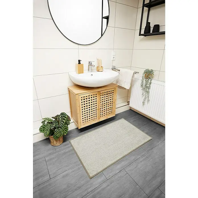 Badteppich Malibu - 60 x 100 cm, Beige, 100% Polyester - Badezimmeransicht mit rechteckiger Badematte aus hellem Stoff vor einem Waschtischunterschrank mit Korbtüren auf grauem Fliesenboden.