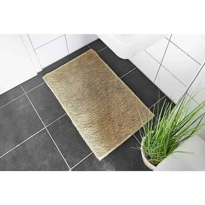 Badteppich Florida - 50 x 80 cm, Taupe, 100% Polyester - Ein rechteckiger, beigefarbener Badteppich liegt auf dunklen Fliesen in einem Badezimmer, neben einem Waschbecken und einer Topfpflanze.