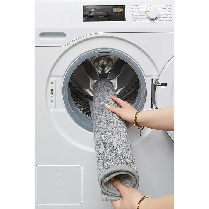 Badteppich Malibu - 60 x 100 cm, Grau, 100% Polyester - Eine Person legt eine graue Badematte in eine weiße Waschmaschine. Die Maschine hat ein digitales Display und verschiedene Waschprogramme.