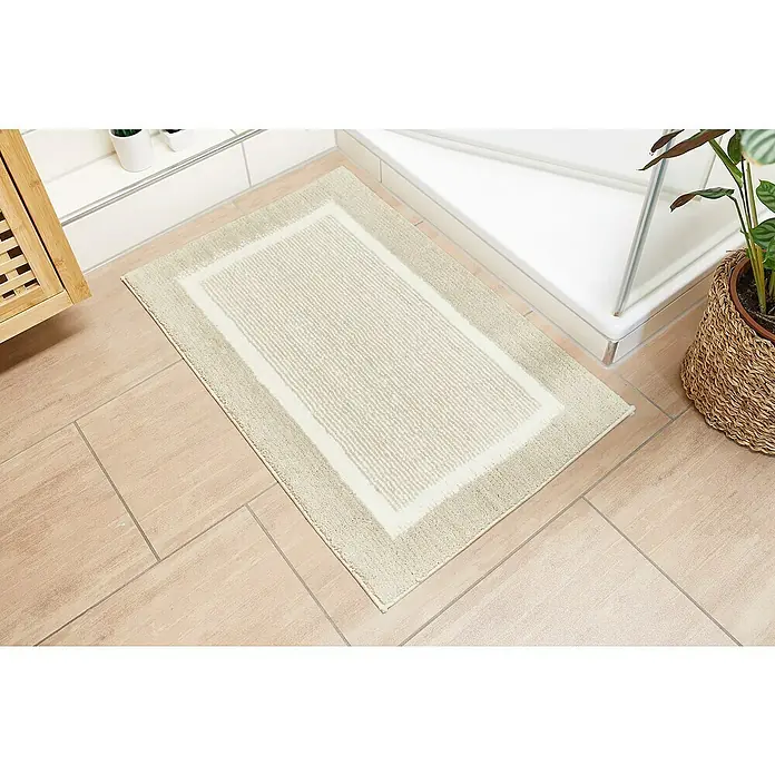 Badteppich Maui - 50 x 80 cm, Beige, 100% Polyester - Ein rechteckiger, beigefarbener Badteppich mit hellem Rand liegt auf hellbraunen Fliesen neben einer Duschkabine und einer Pflanze.
