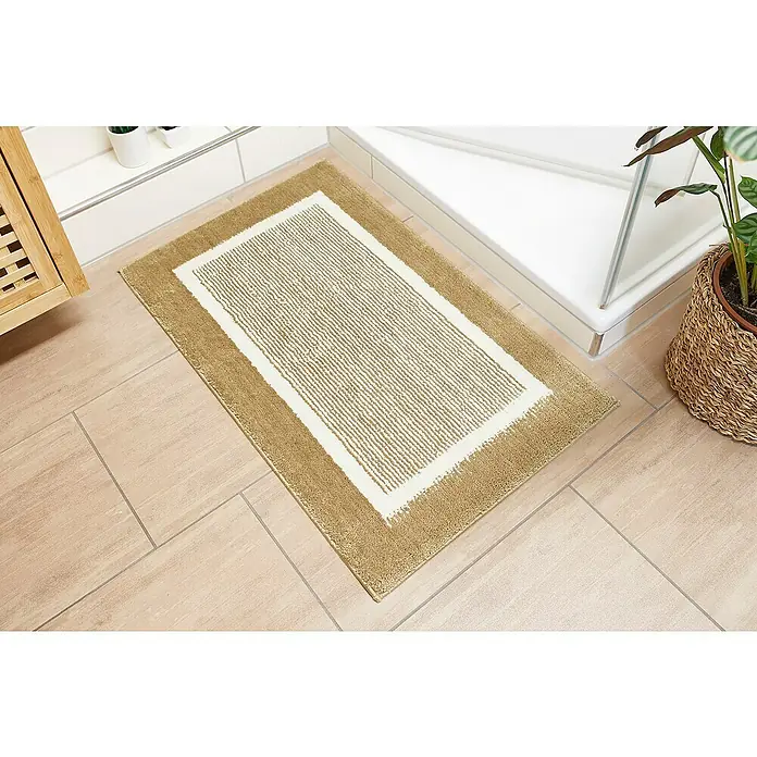 Badteppich Maui - 50 x 80 cm, Taupe, 100% Polyester - Ein rechteckiger, beiger Badezimmerteppich mit weißem Rand liegt auf hellen Fliesen neben einer Dusche und einer Pflanze im Badezimmer.