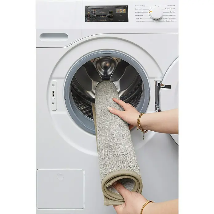 Badteppich Malibu - 60 x 100 cm, Beige, 100% Polyester - Eine Person legt eine graue Badematte in eine weiße Miele-Waschmaschine. Die Maschine zeigt eine Laufzeit von 1:59 an.