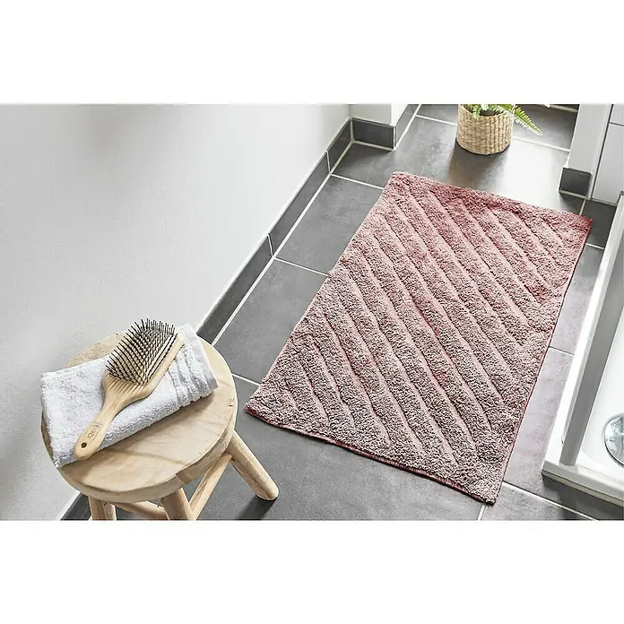 Badteppich Havanna - 50 x 80 cm, Rostrot, Stripe, 80% Baumwolle, 20% Polyester - Ein weicher, rosa Badvorleger mit diagonalem Streifenmuster liegt auf grauen Fliesen neben einem Hocker mit Handtuch und Bürste.