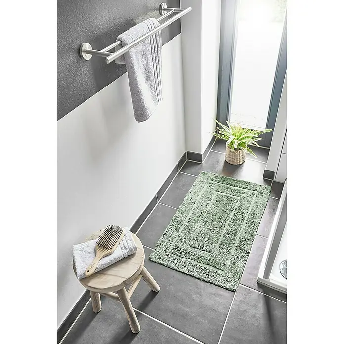 Badteppich Havanna - 50 x 80 cm, Salbei, Border, 80% Baumwolle, 20% Polyester - Ein Badezimmerfoto mit einem grünen Badteppich, Handtüchern auf einer Stange, einem Hocker mit Bürste und Handtuch und einer Zimmerpflanze.