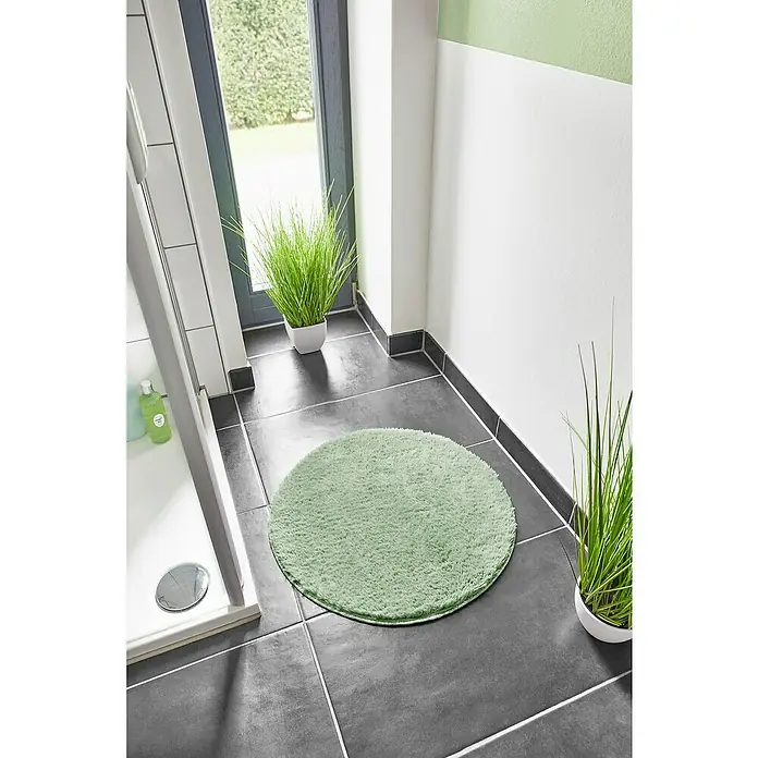 Badteppich Florida - 60 x 60 cm, Salbei, 100% Polyester - Ein runder, hellgrüner Badteppich liegt auf dunklen Fliesen neben einer Dusche und zwei grünen Pflanzen in weißen Töpfen.