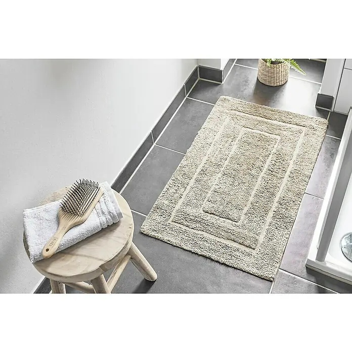 Badteppich Havanna - 50 x 80 cm, Beige, Border, 80% Baumwolle, 20% Polyester - Ein beiger Badteppich mit rechteckigem Design liegt auf grauem Fliesenboden in einem Badezimmer. Auf einem Hocker liegen ein Handtuch und eine Haarbürste.