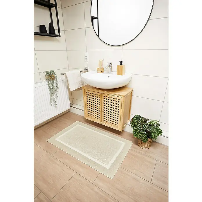 Badteppich Maui - 60 x 100 cm, Beige, 100% Polyester - Ein modernes Badezimmer mit einem hellen Teppich vor einem Waschbeckenunterschrank mit Korbtüren und einem runden Spiegel.