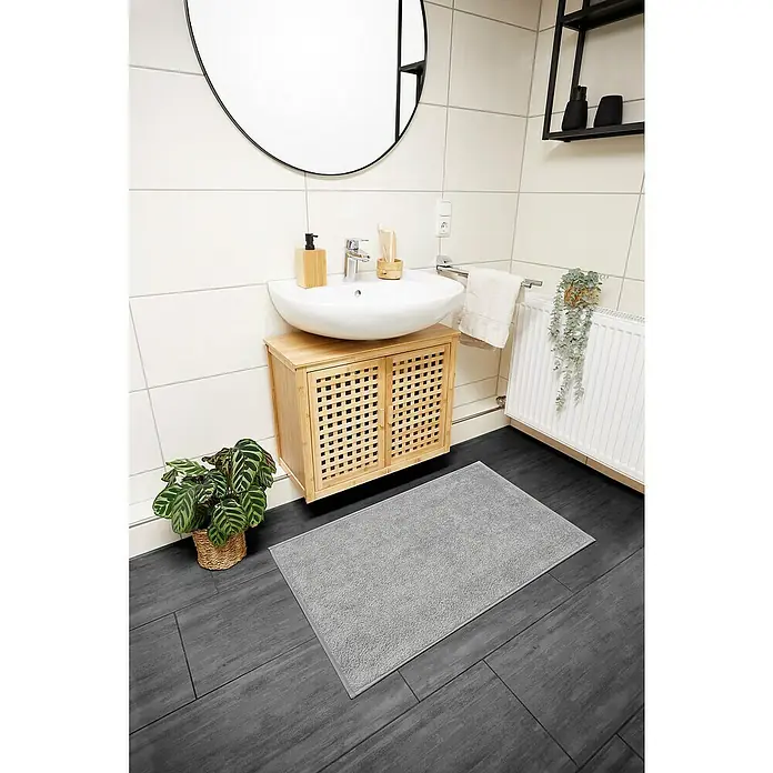 Badteppich Malibu - 60 x 100 cm, Grau, 100% Polyester - Ein hellgrauer, rechteckiger Badteppich liegt auf dunklen Fliesen in einem Badezimmer mit Waschtisch und Dekorationen.