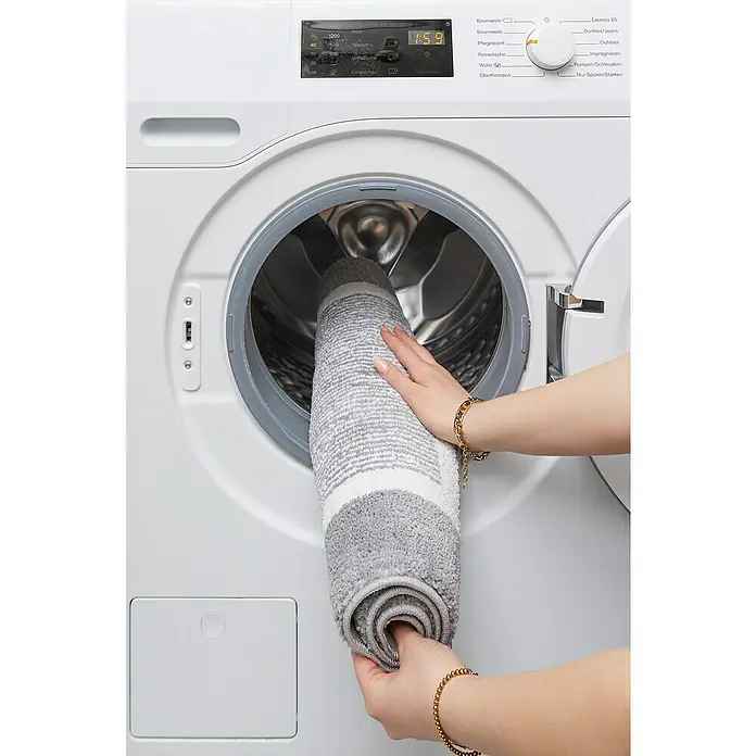 Badteppich Maui - 50 x 80 cm, Grau, 100% Polyester - Eine Person legt einen grau-weiß gestreiften Teppich in eine weiße Waschmaschine.