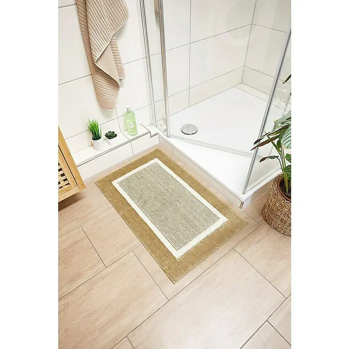 Badteppich Maui - 50 x 80 cm, Taupe, 100% Polyester - Ein rechteckiger, goldfarbener Badezimmerteppich mit weißem Rand liegt vor einer Duschkabine auf hellen Fliesen.