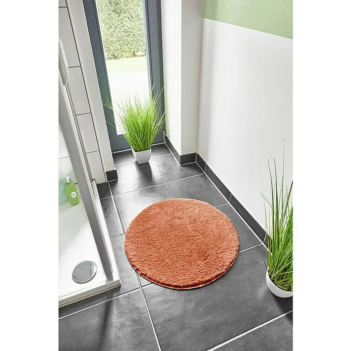 Badteppich Florida - 60 x 60 cm, Rostrot, 100% Polyester - Ein runder, pfirsichfarbener Teppich liegt auf dunklen Fliesen im Badezimmer. Dekorative Pflanzen und eine Duschkabine sind im Hintergrund zu sehen.