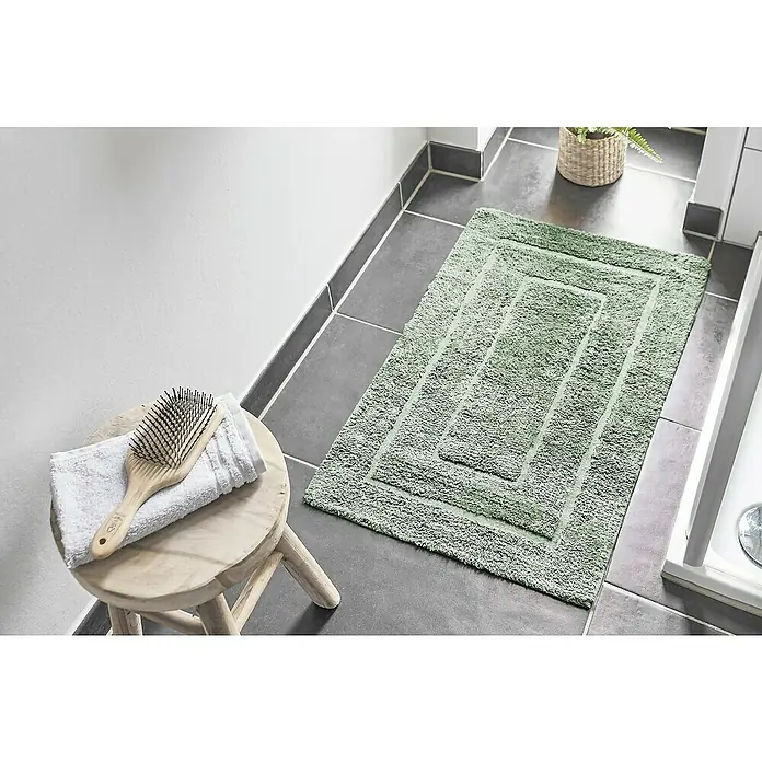 Badteppich Havanna - 50 x 80 cm, Salbei, Border, 80% Baumwolle, 20% Polyester - Ein grüner, rechteckiger Badteppich mit gestuftem Design liegt auf grauen Fliesen, neben einem Hocker mit Handtuch und Bürste.
