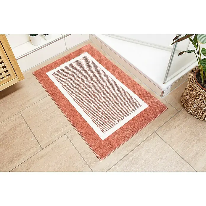 Badteppich Maui - 50 x 80 cm, Rostrot, 100% Polyester - Ein korallenroter Badvorleger mit weißem Rand liegt auf einem hellen, gefliesten Badezimmerboden, neben einer Dusche und einer Holzkommode.