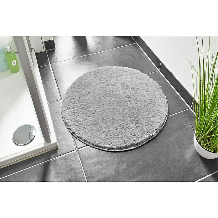 Badteppich Florida - 60 x 60 cm, Grau, 100% Polyester - Ein grauer, runder Badezimmerteppich liegt auf dunklen Fliesen neben einer Dusche und einer Topfpflanze. Der Teppich ist weich und flauschig.