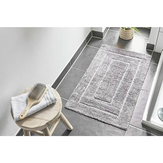 Badteppich Havanna - 50 x 80 cm, Taupe, Border, 80% Baumwolle, 20% Polyester - Ein rechteckiger, flauschiger Badezimmerteppich in Grau liegt auf grauen Fliesen, daneben ein Hocker mit Handtuch und Bürste.