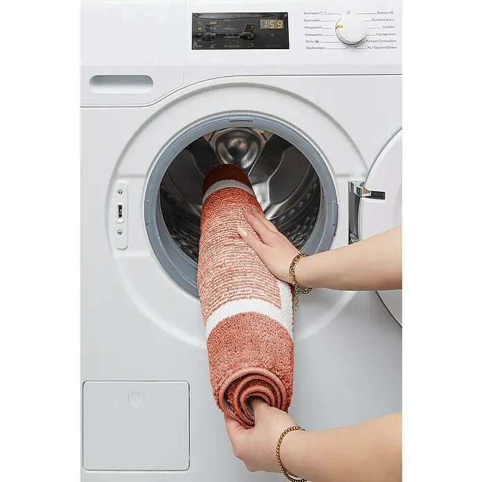 Badteppich Maui - 50 x 80 cm, Rostrot, 100% Polyester - Eine Person legt eine gerollte, orangefarbene Badematte mit weißen Streifen in eine Waschmaschine.