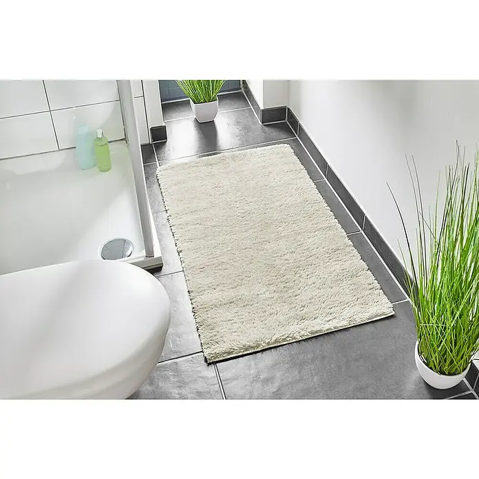 Badteppich Florida - 60 x 100 cm, Beige, 100% Polyester - Ein flauschiger, cremefarbener Teppich liegt auf einem Badezimmerboden mit grauen Fliesen, der für Komfort und Wärme im Bad sorgt.