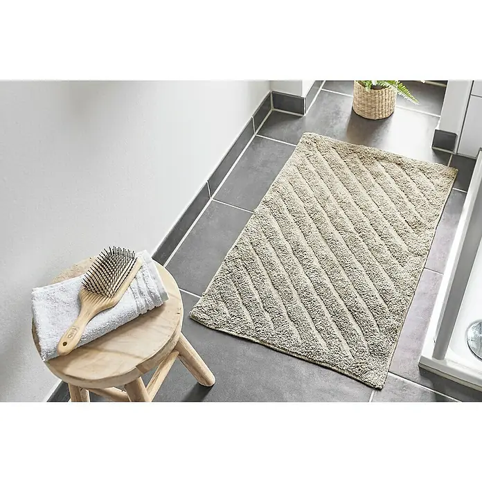 Badteppich Havanna - 50 x 80 cm, Beige, Stripe, 80% Baumwolle, 20% Polyester - Beige Badematte mit diagonalem Streifenmuster auf dunklen Fliesen, neben einem Hocker mit Handtuch und Bürste.