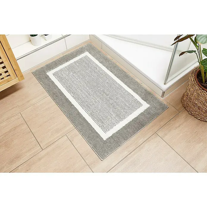 Badteppich Maui - 50 x 80 cm, Grau, 100% Polyester - Ein rechteckiger Badteppich mit grauem Rand und weiß-grauem Innenbereich liegt auf einem beigen Fliesenboden.