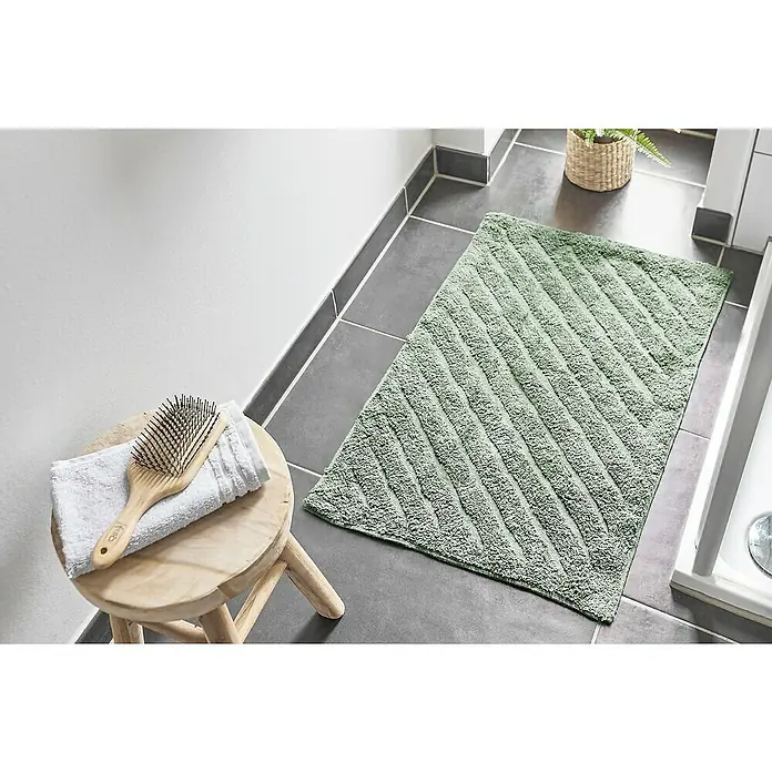 Badteppich Havanna - 50 x 80 cm, Salbei, Stripe, 80% Baumwolle, 20% Polyester - Ein grüner Badteppich mit Streifenmuster liegt auf grauen Fliesen in einem Badezimmer. Auf einem Hocker liegen eine Bürste und ein weißes Handtuch.