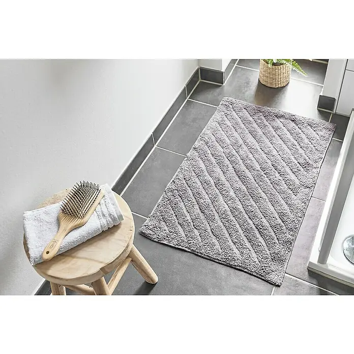 Badteppich Havanna - 50 x 80 cm, Taupe, Stripe, 80% Baumwolle, 20% Polyester - Ein grauer Badteppich mit diagonalem Streifenmuster liegt auf grau gefliestem Boden, neben einem kleinen Hocker mit Handtuch und Bürste.