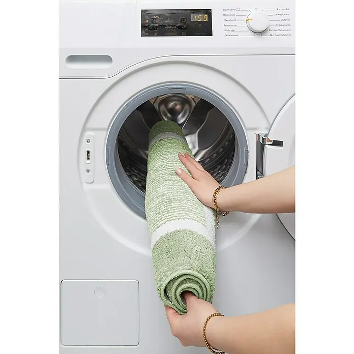 Badteppich Maui - 60 x 100 cm, Salbei, 100% Polyester - Eine Person legt eine hellgrüne Badematte mit weißen Streifen in eine weiße Miele Waschmaschine.