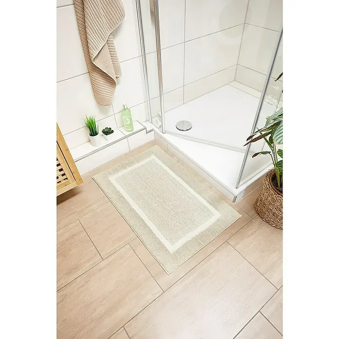Badteppich Maui - 50 x 80 cm, Beige, 100% Polyester - Beiger Badteppich mit Rahmen auf Fliesenboden vor einer Dusche mit Topfpflanze und Handtuch.