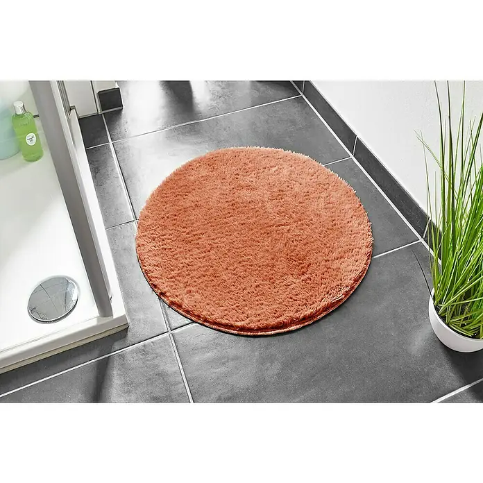 Badteppich Florida - 60 x 60 cm, Rostrot, 100% Polyester - Ein runder, pfirsichfarbener Badteppich liegt auf dunklen Fliesen, neben einer Dusche und einer Zimmerpflanze. Perfekt für ein stilvolles Badezimmer.