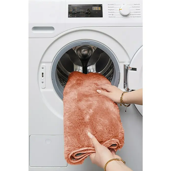 Badteppich Florida - 50 x 80 cm, Rostrot, 100% Polyester - Nahaufnahme einer Person, die einen flauschigen, lachsfarbenen Teppich in eine weiße Waschmaschine legt.