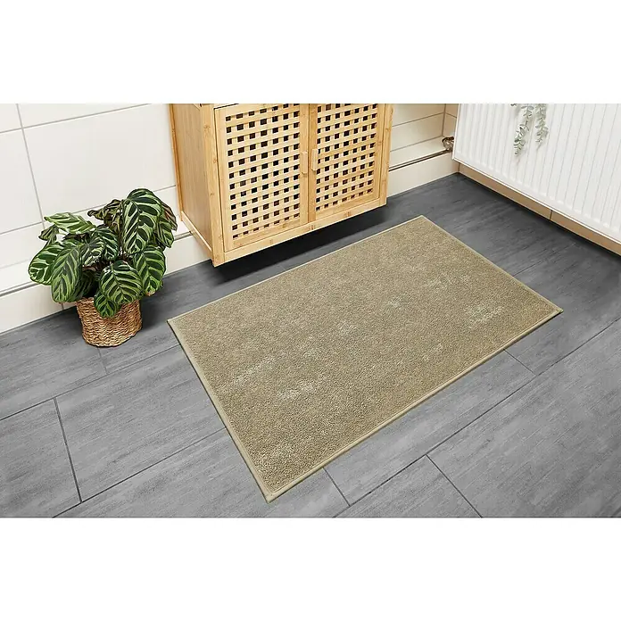 Badteppich Malibu - 60 x 100 cm, Taupe, 100% Polyester - Beige Badematte auf grauem Fliesenboden vor einem Holzschrank und einer Zimmerpflanze. Sie sorgt für Komfort und Stil im Badezimmer.