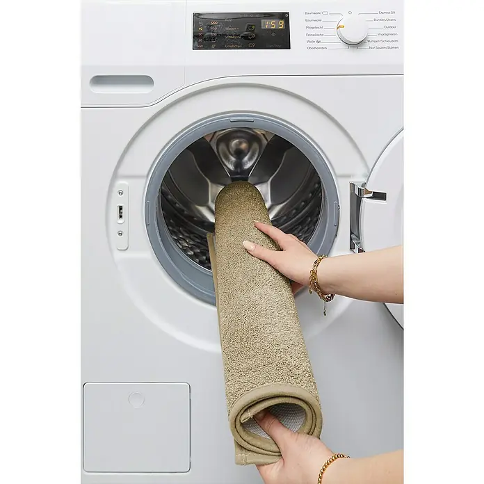 Badteppich Malibu - 60 x 100 cm, Taupe, 100% Polyester - Eine Person legt eine gerollte, hellbraune Badematte in eine weiße Waschmaschine mit geöffneter Tür.