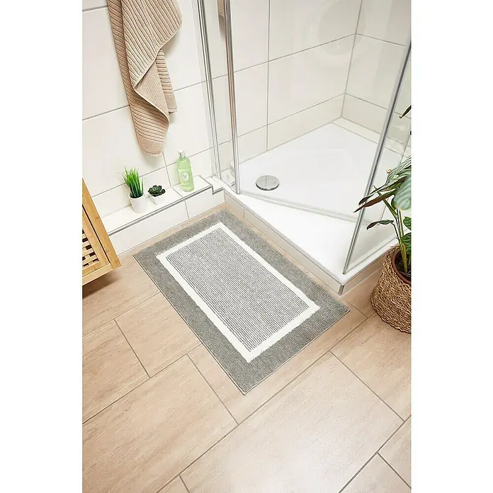 Badteppich Maui - 50 x 80 cm, Grau, 100% Polyester - Ein rechteckiger, grauer Badteppich mit weißem Rand liegt auf hellbraunen Fliesen vor einer Dusche.