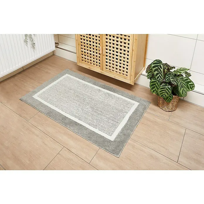 Badteppich Maui - 60 x 100 cm, Grau, 100% Polyester - Ein rechteckiger, grauer Badezimmerteppich mit weißem Rand liegt auf dem Boden vor einem Holzschrank und einer Pflanze.
