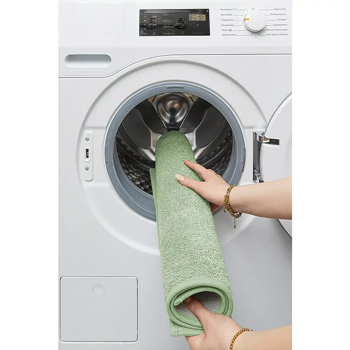 Badteppich Malibu - 60 x 100 cm, Salbei, 100% Polyester - Eine Person legt eine grüne Badematte in eine weiße Waschmaschine mit geöffneter Tür.