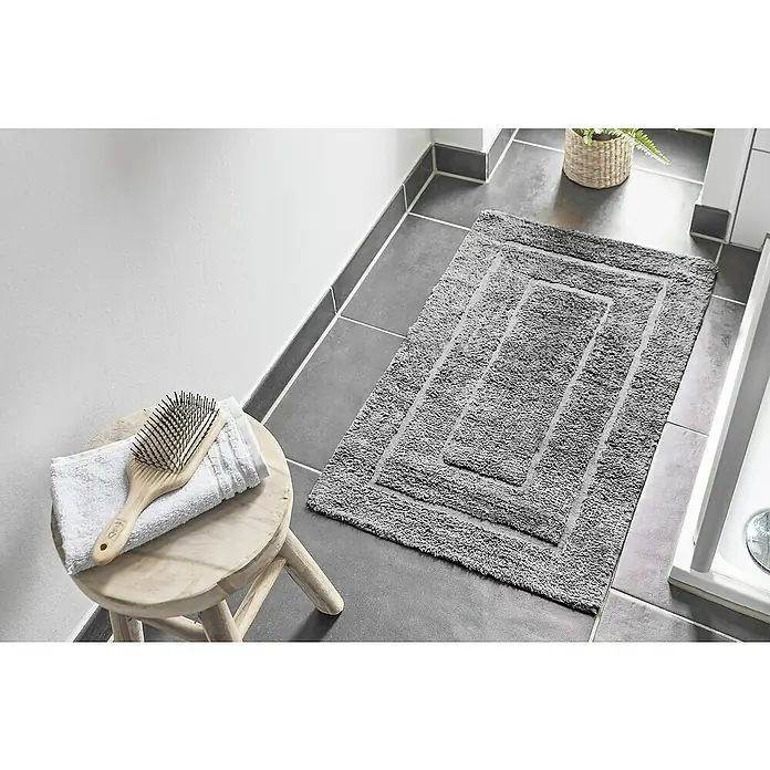 Badteppich Havanna - 50 x 80 cm, Grau, Border, 80% Baumwolle, 20% Polyester - Bild eines grauen Badezimmerteppichs mit strukturierter Oberfläche, der auf dunklen Fliesen liegt, neben einem Hocker mit Handtuch und Bürste.