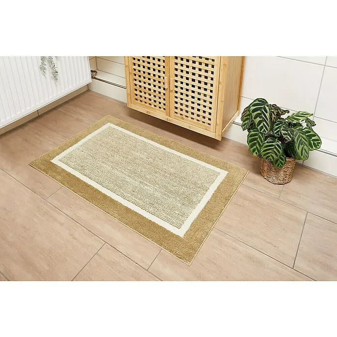 Badteppich Maui - 60 x 100 cm, Taupe, 100% Polyester - Ein rechteckiger, beigefarbener Badteppich mit einem cremefarbenen Innenbereich liegt auf einem hellen Holzboden. Daneben steht ein Schrank und eine Pflanze.