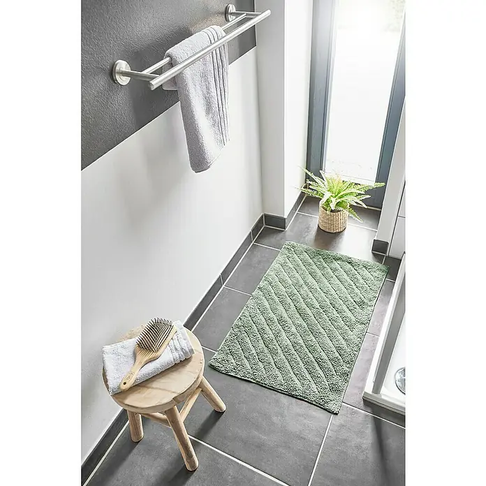 Badteppich Havanna - 50 x 80 cm, Salbei, Stripe, 80% Baumwolle, 20% Polyester - Dieses Badezimmerbild zeigt eine grüne Badematte mit diagonalem Muster auf einem dunklen Fliesenboden, neben einem Hocker mit Handtüchern.