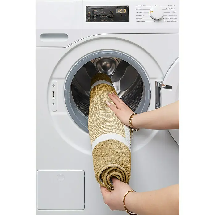Badteppich Maui - 50 x 80 cm, Taupe, 100% Polyester - Eine Person legt eine gerollte, gestreifte Badematte in eine weiße Waschmaschine, um sie zu reinigen.