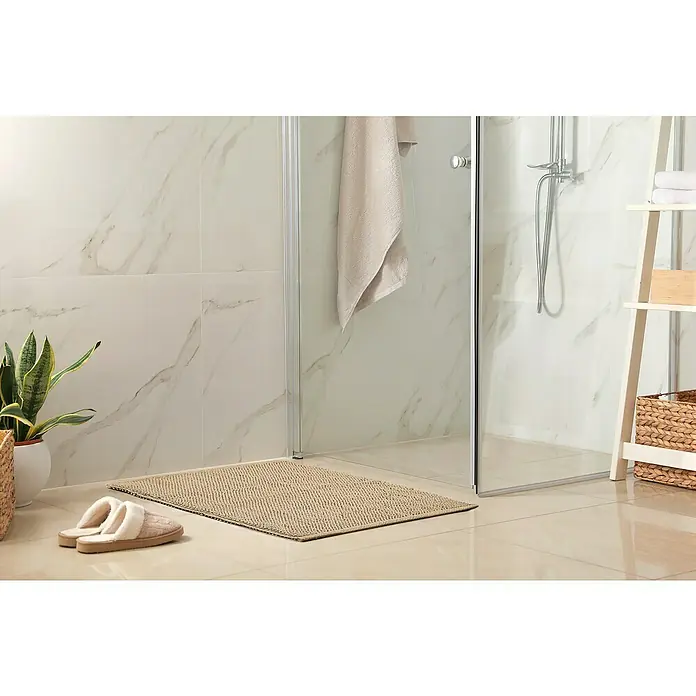 Badteppich Atlanta - 50 x 80 cm, Beige, 100% Baumwolle - Beige Badematte liegt vor einer Dusche mit Glastüren, neben Hausschuhen und einer Topfpflanze. Ideal für ein stilvolles Badezimmer.