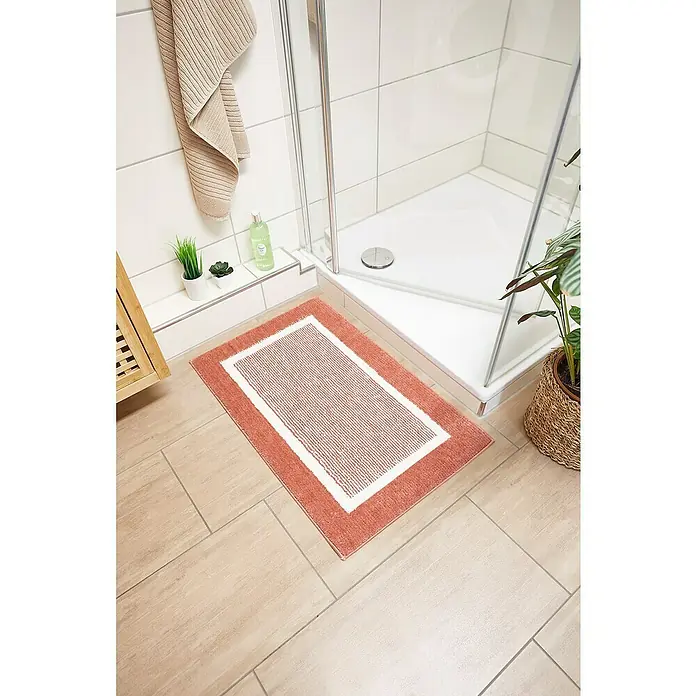 Badteppich Maui - 50 x 80 cm, Rostrot, 100% Polyester - Ein rechteckiger, korallenroter Badteppich mit weißem Rand liegt vor einer Dusche in einem Badezimmer mit Fliesenboden.