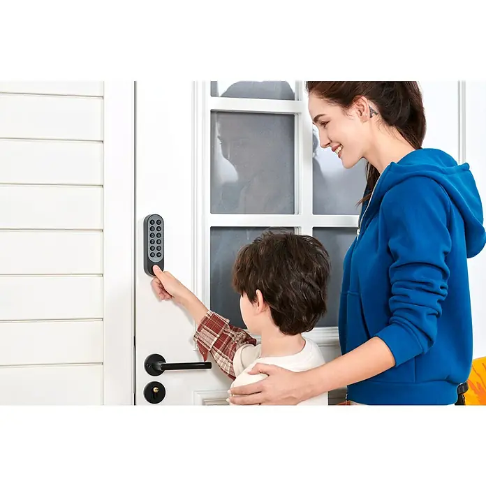 Aqara Elektronisches Türschloss SMART LOCK U200 - Bedienung: Fingerabdruck, Zugangscode, Smartphone, Geeignet für: Türen, Schwarz - Ein Kind, das von einer Frau begleitet wird, bedient ein elektronisches Türschloss mit Tastenfeld an einer weißen Tür.