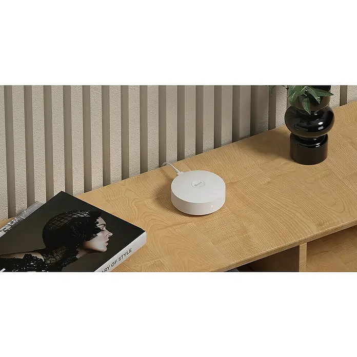EZVIZ Steuerzentrale A3-R200 - Weiß, L x B x H: 91 x 102 x 107 mm - Nahaufnahme eines runden, weißen EZVIZ-Geräts auf einem Holzregal mit einem Kabel und einem Buch.