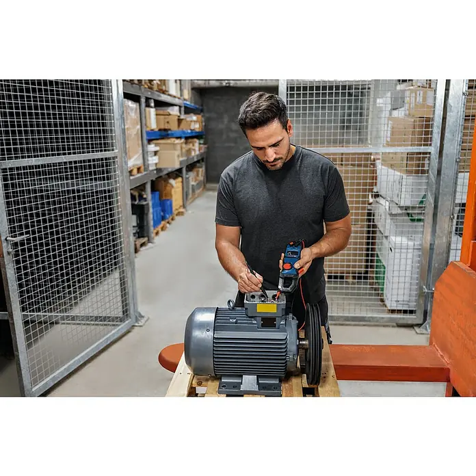 Bosch Professional Stromzange GMC 600-15 - 600 V, Blau, Schwarz - Mann prüft einen grauen Elektromotor mit einem blauen Messgerät auf einer Palette in einem Lagerhaus mit Regalen und Metallgitterkäfigen.