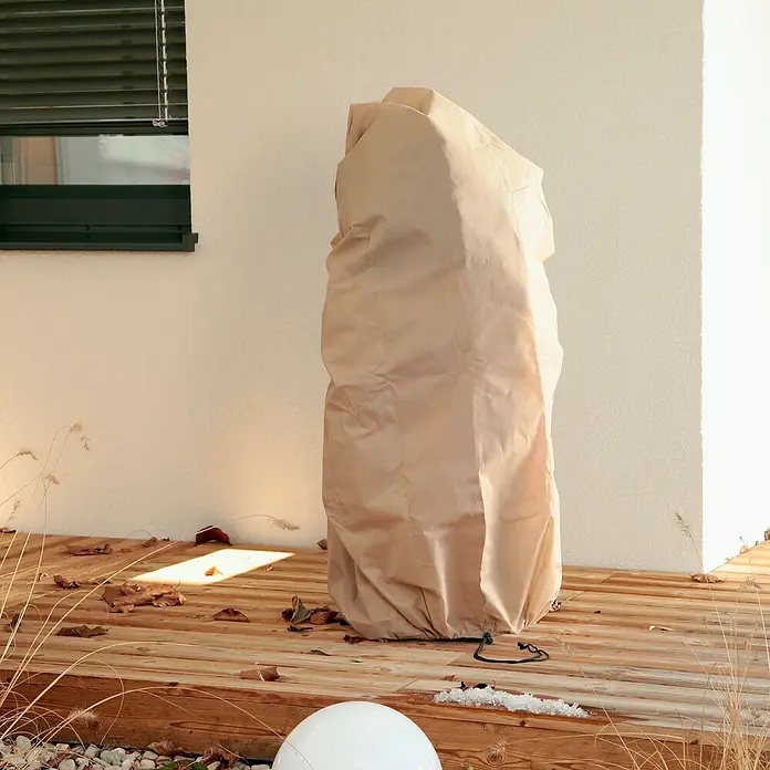 Windhager Winterschutzhaube Superprotect L - Ø x H: 60 x 100 cm, 70 g/m² - Ein winterfester Pflanzensack schützt eine Pflanze vor Frost, der auf einer Terrasse steht. Der Sack ist beige und aus einem robusten Material.